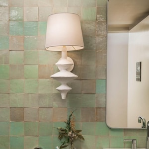 May include: A white wall-mounted lamp with a cream lampshade. The lamp has a distinctive, layered design and is fixed to a wall with square, green tiles. A mirror and a small vase of flowers are also visible.