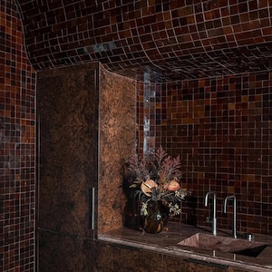 May include: A bathroom with brown mosaic tile walls and ceiling. A dark brown cabinet is on the left, and a sink with silver faucets is on the right. A vase of dried flowers sits on the countertop.