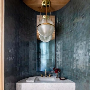 Op de afbeelding: Een moderne badkamer met een gebogen muur betegeld met turquoise vierkanten. Een grote spiegel weerspiegelt een bolvormige glazen lamp met een goudkleurig frame. De wastafel heeft goudkleurige armaturen en een wit aanrechtblad.