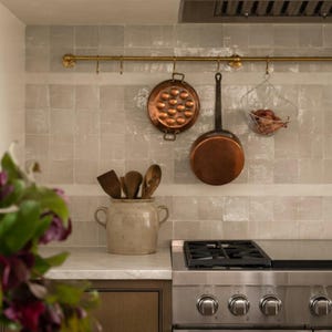 Könnte beinhalten: Eine Küchenszene mit Kupferkochgeschirr, einem Drahtkorb und Holzutensilien. Eine Messingstange hält die hängenden Gegenstände vor einer hellgrauen gefliesten Rückwand. Ein Keramiktopf enthält Holzlöffel und -spatel. Ein Edelstahl-Gasherd befindet sich im Vordergrund.