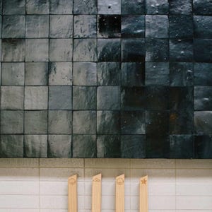 May include: A wall of dark grey square tiles with a glossy finish. Below, four wooden utensils with a circular design are arranged against a white tiled surface. The tiles create a textured, geometric pattern.