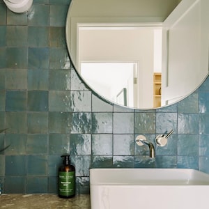 May include: A modern bathroom with teal square tiles, a white rectangular sink, and a round mirror. A chrome faucet and a dark brown bottle of eucalyptus mint soap are visible. The mirror reflects a doorway and a glimpse of another room.