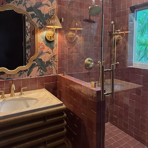 May include: A bathroom featuring a red-tiled shower with gold fixtures and a glass door. A vanity with a gold-framed mirror and floral wallpaper is visible. The room includes a window offering a view of green foliage.