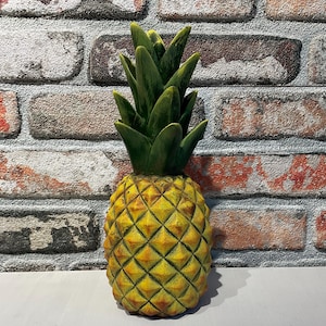 May include: A decorative pineapple sculpture with a textured yellow body and green leafy top. The pineapple is set against a brick wall background. The sculpture is a unique home decor item.