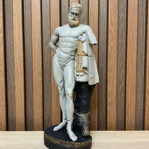 Könnte beinhalten: Graue Statue einer muskulösen männlichen Figur mit Bart, drapiert mit einem weißen Tuch mit goldenen Akzenten. Die Statue hat einen fehlenden Arm und steht auf einem schwarzen Sockel mit goldenen Verzierungen. Dekorationsartikel.