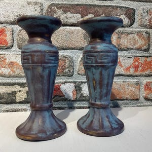 K&ouml;nnte beinhalten: Zwei Kerzenhalter im antiken Stil mit einem verwitterten blau-braunen Finish. Die Kerzenhalter haben einen ausgestellten Sockel, einen detaillierten Mittelteil und eine breite Oberseite. Der Hintergrund ist eine Backsteinmauer.