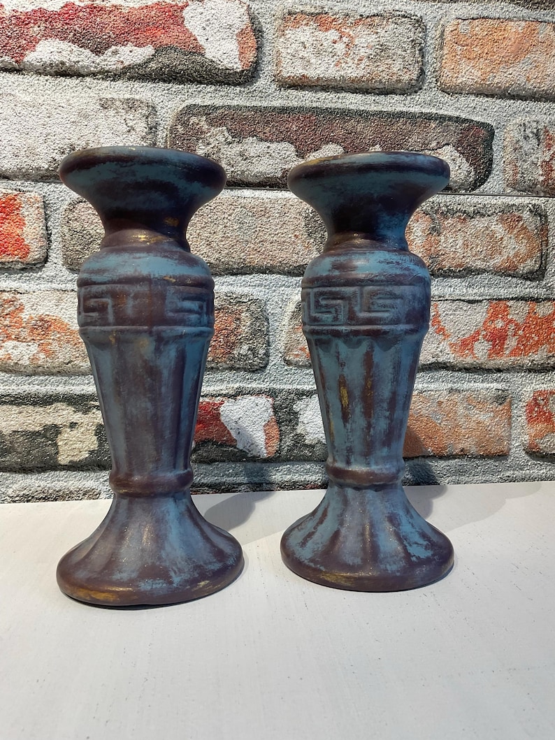 K&ouml;nnte beinhalten: Zwei Kerzenhalter im antiken Stil mit einem verwitterten blau-braunen Finish. Die Kerzenhalter haben einen ausgestellten Sockel, einen detaillierten Mittelteil und eine breite Oberseite. Der Hintergrund ist eine Backsteinmauer.