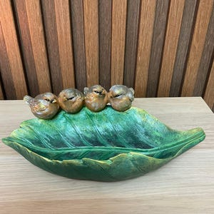 May include: A decorative leaf-shaped dish with four small bird figurines. The dish is painted green with gold accents, and the birds are bronze-colored. The leaf dish is a decorative item.