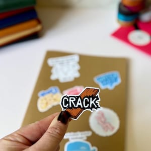 Peut inclure: Un autocollant avec le mot "CRACK" en contour noir, avec un motif de planche de bois marron. L'autocollant est tenu dans une main, avec d'autres autocollants sur un cahier marron en arrière-plan.