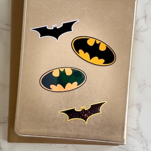 Puede incluir: Un cuaderno rectangular de color dorado con cuatro pegatinas con temática de Batman. Las pegatinas incluyen una silueta de murciélago negra, un logotipo ovalado clásico amarillo y negro, un logotipo ovalado verde oscuro y dorado, y un murciélago negro con estrellas.