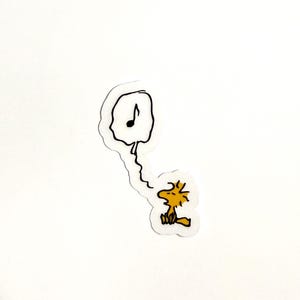 May include: A die-cut sticker featuring a yellow cartoon bird character sitting and looking up at a speech bubble containing a single black musical note. The design is outlined in black ink on a white background with a thin white border.