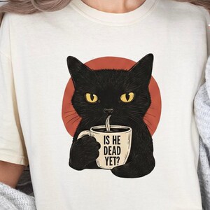 May include: Cream-colored t-shirt featuring a black cat holding a coffee mug. The cat is set against a red circle, with the text "IS HE DEAD YET?" on the mug. The cat has yellow eyes.