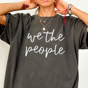 May include: Dark gray oversized t-shirt with the white text "we the people". The shirt has short sleeves and a relaxed fit. The shirt is made of a soft, comfortable material.