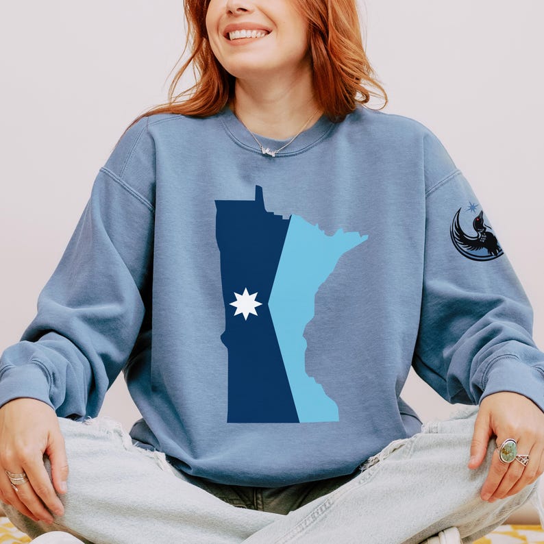 May include: A blue sweatshirt featuring a graphic of the state of Minnesota in shades of blue, with a white starburst design. The sweatshirt has a crew neck and long sleeves. A black graphic of a bird and crescent moon is on the upper sleeve.