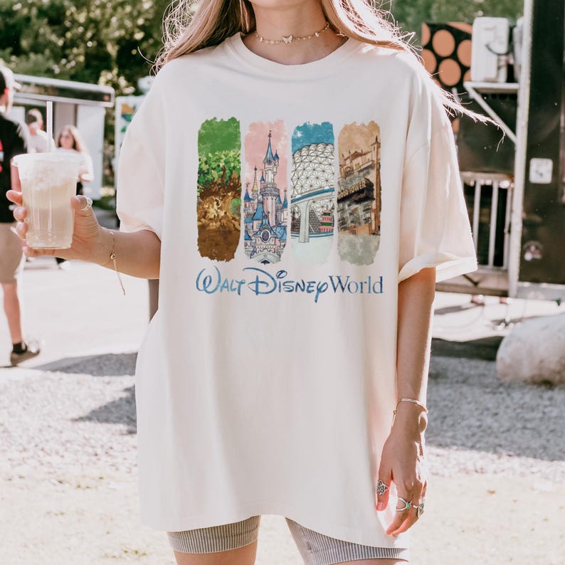 Puede incluir: Camiseta color crema con un gr&aacute;fico colorido de cuatro paneles rectangulares con im&aacute;genes de Disney World. Los paneles incluyen un castillo, un &aacute;rbol y otros elementos arquitect&oacute;nicos. Las palabras "Walt Disney World" est&aacute;n impresas en azul debajo.