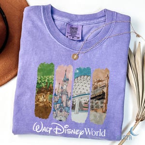 Puede incluir: Una camiseta lavanda Comfort Colors con cuatro gr&aacute;ficos rectangulares verticales que representan monumentos de Walt Disney World. Las palabras "Walt Disney World" est&aacute;n impresas en blanco debajo. Un collar dorado est&aacute; sobre la camiseta.