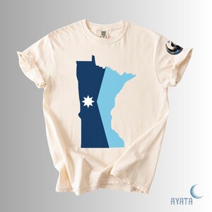 May include: Cream-colored t-shirt featuring a stylized map of Minnesota in navy blue and light blue, with a white starburst design. The shirt has rolled-up sleeves and a small graphic on the right sleeve.