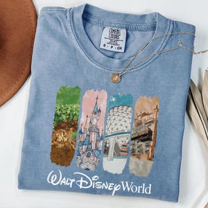 Puede incluir: Una camiseta Comfort Colors azul claro con un gr&aacute;fico de cuatro rect&aacute;ngulos coloridos con im&aacute;genes de Disney World. Las palabras "Walt Disney World" est&aacute;n impresas en blanco debajo. Un collar dorado con un colgante descansa sobre la camiseta.