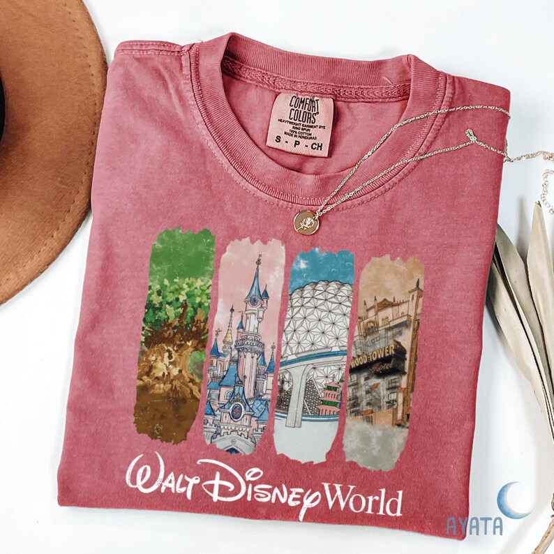 Puede incluir: Una camiseta Comfort Colors de color rosa rojizo con un gr&aacute;fico de los monumentos de Walt Disney World. La camiseta tiene cuello redondo y mangas cortas. Un collar plateado con un peque&ntilde;o colgante est&aacute; drapeado sobre la camiseta. Las palabras "Walt Disney World" est&aacute;n impresas en la parte inferior.