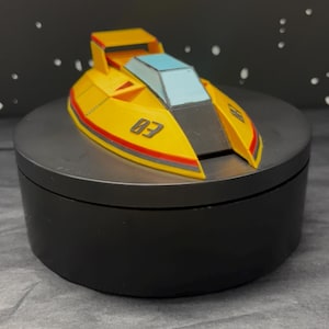 Op de afbeelding: Een gele speelgoedboot met een rode en zwarte streep en het nummer "03" aan de zijkant. De boot heeft een blauwe voorruit en staat op een zwart platform.