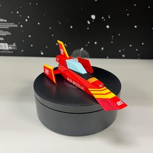 May include: A red and yellow toy spaceship with the text "RED GAZELLE" and the number "01" printed on it. The spaceship has a blue cockpit and is displayed on a black rotating stand.