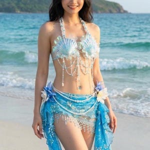 May include: A mermaid-themed costume featuring a seashell bra and a blue skirt with pearl and crystal accents. The bra is adorned with light blue seashells and delicate white lace. The skirt is a flowing blue fabric with a pearl belt and crystal fringe.