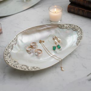 Puede incluir: Una bandeja ovalada de joyería de nácar con un borde decorativo plateado. La bandeja contiene varios anillos, pendientes y un collar. Una vela encendida y un espejo están en el fondo.