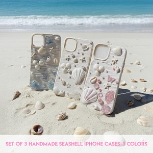 May include: Three handmade seashell iPhone cases in white, adorned with seashells, starfish, and pearls. The cases are displayed on a sandy beach with the sea in the background. The text at the bottom reads: SET OF 3 HANDMADE SEASHELL IPHONE CASES - 3 COLORS.