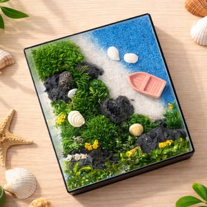 Pode incluir: Uma cena de praia em miniatura numa caixa com moldura preta. A cena inclui musgo verde, rochas pretas, areia branca, glitter azul e branco, um pequeno barco rosa e conchas. Flores amarelas adicionam um toque de cor.