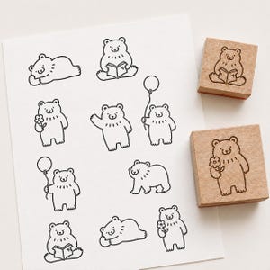 Può includere: Due timbri in gomma quadrati in legno con un'illustrazione a linee di un orso che tiene un fiore. Un foglio di carta bianco mostra diverse impronte a inchiostro nero di orsi in varie pose, tra cui intenti a leggere, con palloncini e che salutano.