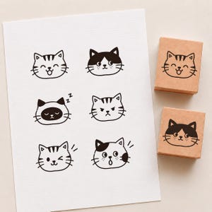 Può includere: Un foglio di carta bianca con sei distinte illustrazioni a inchiostro nero di volti di gatto con varie espressioni, accompagnate da due timbri in gomma quadrati in legno, ognuno dei quali mostra uno dei disegni di gatto in nero.