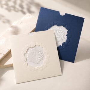 May include: Two square envelopes, one in cream and one in deep navy blue, each featuring a central embossed scallop shell design surrounded by a circular border of various smaller seashells and a subtle wave pattern along the edges.