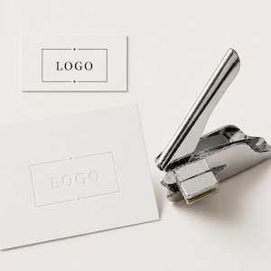 Puede incluir: Una herramienta de estampado manual de metal plateado junto a dos tarjetas rectangulares blancas. Una tarjeta presenta un marco rectangular impreso en negro con el texto "LOGO" dentro, mientras que la otra muestra un diseño de logo en relieve.