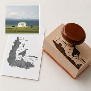 May include: A rectangular wooden rubber stamp with a dark brown circular handle. The stamp features a black ink illustration of a house near a body of water, surrounded by trees. A white card displays the same black ink house illustration.