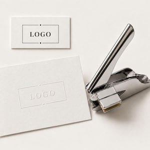 May include: A silver metal hand embosser tool positioned next to two white rectangular cards. One card features a printed black rectangular frame with the text "LOGO" inside, while the other card displays the same design as a blind embossed impression.
