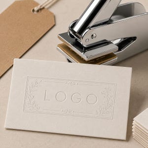 Könnte beinhalten: Eine cremefarbene Visitenkarte mit dem Wort „LOGO“ in der Mitte geprägt, umrahmt von floralen Designs. Eine silberne Metallprägemaschine befindet sich über der Karte und ist bereit, einen erhabenen Eindruck zu erzeugen. Ein braunes Etikett befindet sich im Hintergrund.