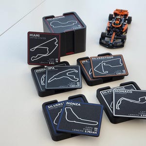 May include: A collection of square black coasters featuring white outlines of Formula 1 race tracks, including Miami, Spa, Silverstone, and Monza, with text detailing laps, turns, and length in km. A small orange and black toy race car is included.