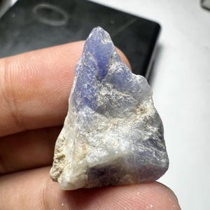 Puede incluir: Un cristal de tanzanita en bruto de forma triangular que presenta un degradado de tonos azul violeta translúcidos que se transforman en depósitos minerales blancos y grises opacos y texturizados en su superficie irregular y ápice puntiagudo.