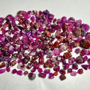 Puede incluir: Una gran colección de piedras preciosas de rubí en bruto y sin pulir en varios tonos de rosa intenso y magenta. Estos pequeños fragmentos minerales irregulares presentan texturas naturales, superficies de translúcidas a opacas e inclusiones blancas o oscuras.