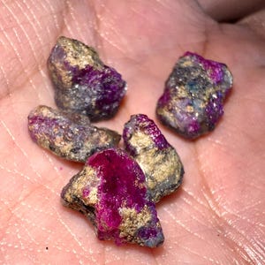 以下が含まれることがあります： いくつかのラフカットのルビーの宝石のクローズアップ。石は、金とグレーの領域を持つ鮮やかなマゼンタ色を示しています。宝石は不規則な形をしており、自然で未研磨の標本のようです。