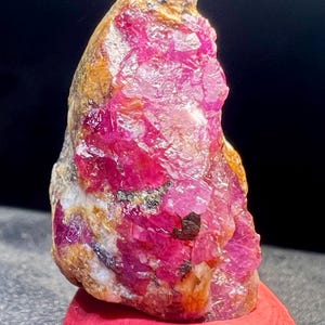 Peut inclure: Un spécimen minéral brut et non poli présentant des structures cristallines magenta vif et rose foncé incrustées dans une matrice rocheuse texturée de couleur beige et brune, avec des facettes naturelles et des surfaces réfléchissantes.