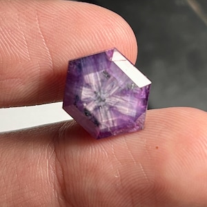 Puede incluir: Una gema facetada, hexagonal, de color púrpura intenso. La gema presenta un patrón en forma de estrella en su interior, con una mezcla de tonos claros y oscuros. La piedra se sostiene entre los dedos, mostrando su tamaño y detalle.