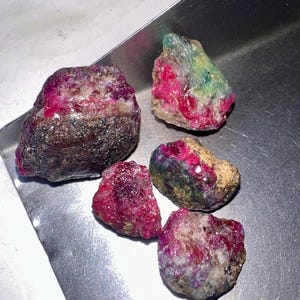 May include: Five rough gemstones with a mix of colors, including deep reds, pinks, greens, and browns. The stones have a natural, unpolished appearance, with visible textures and variations in color. The gemstones are displayed on a metallic surface.