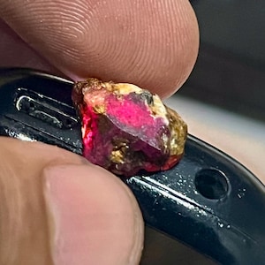 Puede incluir: Una gema en bruto con un vibrante tono rosa y rojo, acentuada por tonos dorados y marrones. El mineral en bruto se sostiene contra una superficie negra, mostrando su textura natural y sin pulir. Esta imagen resalta la belleza de una gema natural.