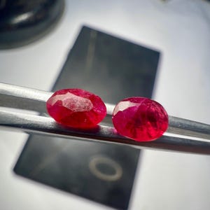 Może przedstawiać: Dwa owalne, żywe czerwone kamienie szlachetne są trzymane przez srebrną pęsetę. Kamienie są umieszczone na rozmytym tle, sugerując kontekst biżuterii lub oprawy kamieni szlachetnych. Kamienie są fasetowane, odbijając światło.