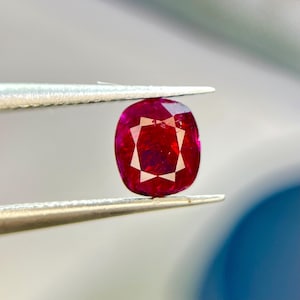May include: A vibrant, cushion-cut ruby gemstone with a deep red hue and multifaceted surface, held securely between the tips of a pair of silver-toned metal tweezers. The stone displays internal light reflections and a clear, polished finish.