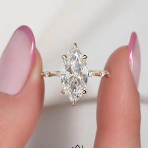 May include: A marquise-cut solitaire ring featuring a clear, faceted center stone set in a yellow gold band with small accent stones along the shank. The ring is positioned centrally, and the text OM GEMS JEWEL is printed at the bottom.