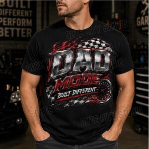 Op de afbeelding: Zwart T-shirt met een grafisch ontwerp met de woorden "DAD MODE" in een metallic zilveren en rode lettertype. Het ontwerp bevat een geblokte vlag, vlammen en een snelheidsmeter. De tekst "BUILT DIFFERENT" is ook opgenomen.