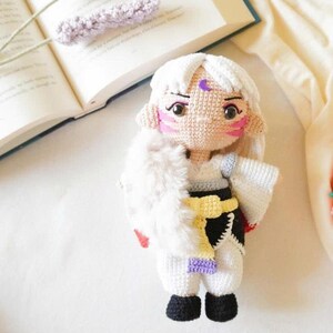 Puede incluir: Muñeca de ganchillo hecha a mano con cabello blanco, una luna creciente púrpura en la frente y marcas rosadas en las mejillas. La muñeca viste un atuendo blanco con detalles negros, una faja amarilla y botas negras.