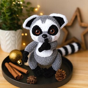 May include: A handmade gray, white, and black crochet lemur plush toy with large brown eyes and a striped tail. The lemur is sitting on a black tray with cinnamon sticks, a gold ornament, and a pine cone. The lemur has its finger to its lips.